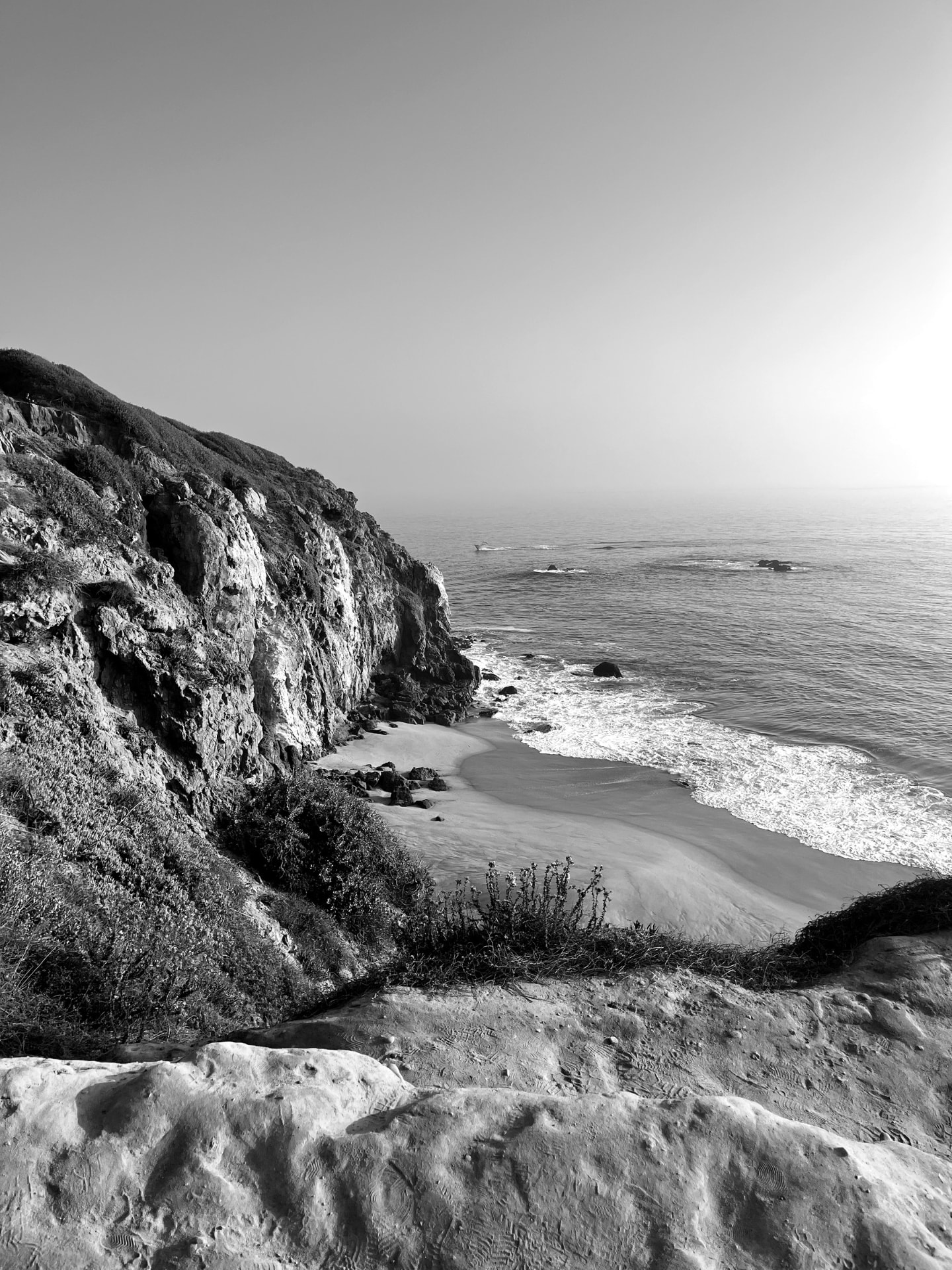 A coastal cliff dropping to a small beach, in black and white